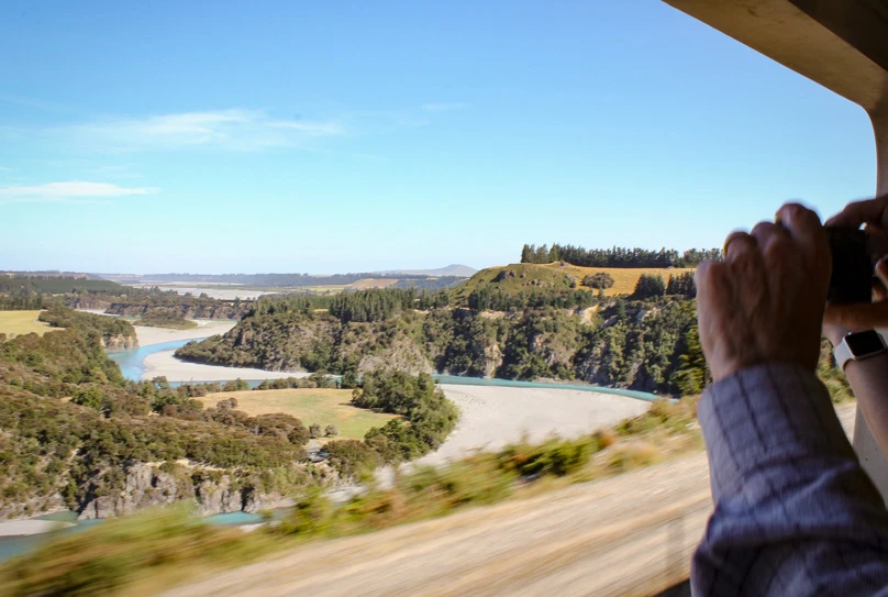 Person taking a photo out the open-air viewing carriage on the TranzAlpine passing Waimakariri River